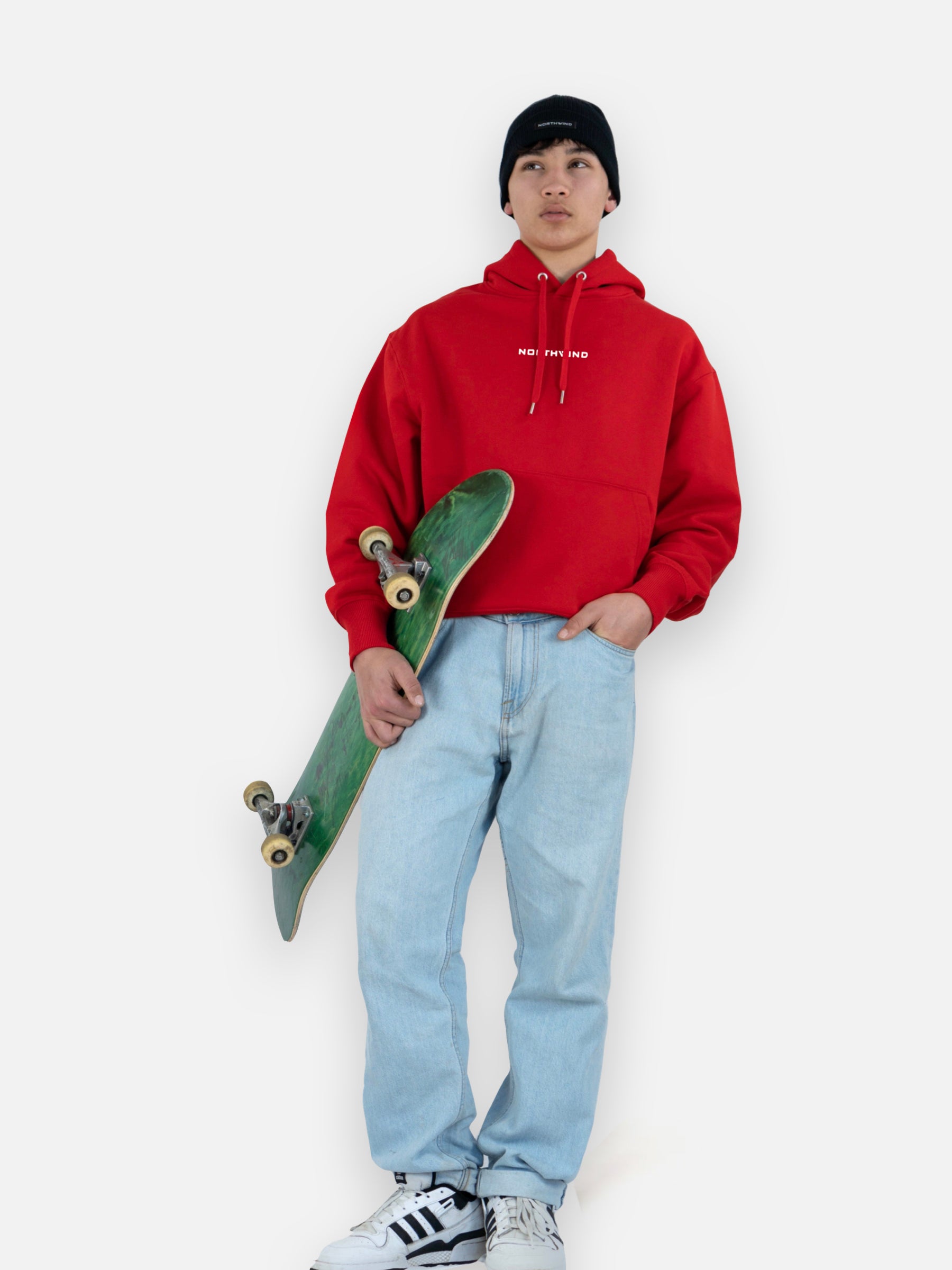 Man wearing a black organic cotton beanie with a Northwind logo, and a red organic cotton oversize heavy Northwind hoodie with embroidered logo on the chest, holding a skate board
